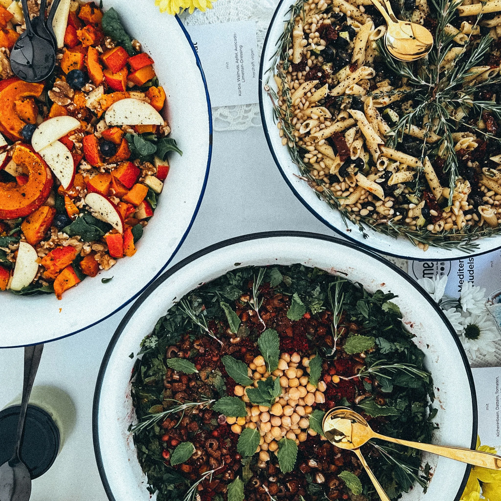 Drei große Salatschüsseln von oben fotografiert. Oben links ein bunter Salat mit Kürbis, Apfel, Blaubeeren und Walnüssen. Oben rechts ein Nudel-Pinienkern-Salat mit Kräutern. Unten ein Salat mit Kichererbsen, Minze und weiteren grünen Blättern.