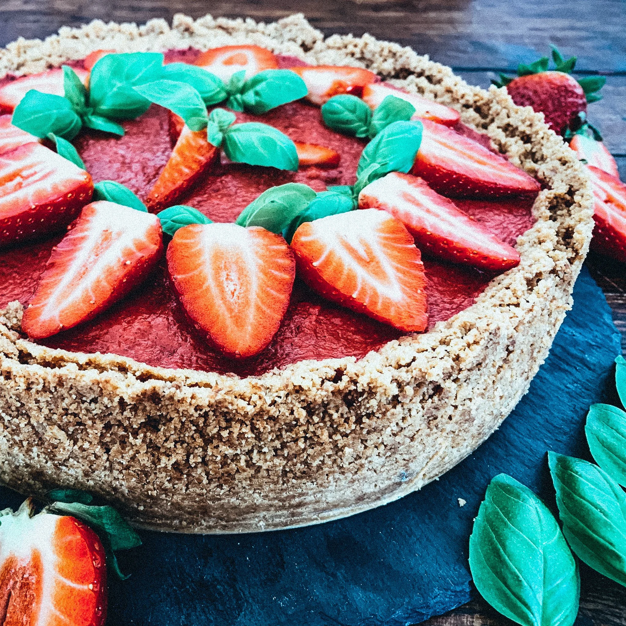 Erdbeerkuchen mit frischen Erdbeerscheiben und Basilikumblättern dekoriert, auf einem dunklen Schieferuntergrund. Der Kuchen hat einen knusprigen Keksboden.