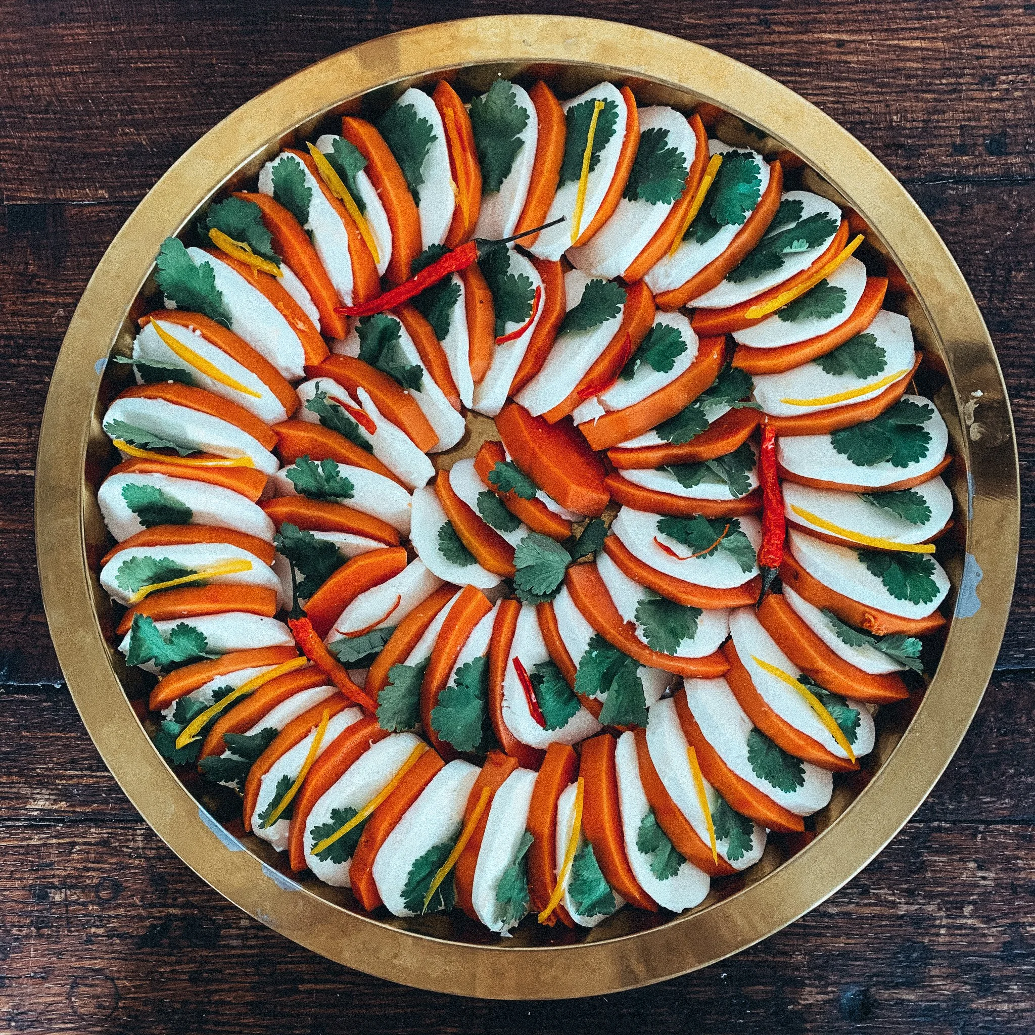 Ein rundes Tablett mit kunstvoll arrangiertem Fingerfood, bestehend aus weißen Happen, Orangenscheiben, Koriander und roten Chili-Schoten.