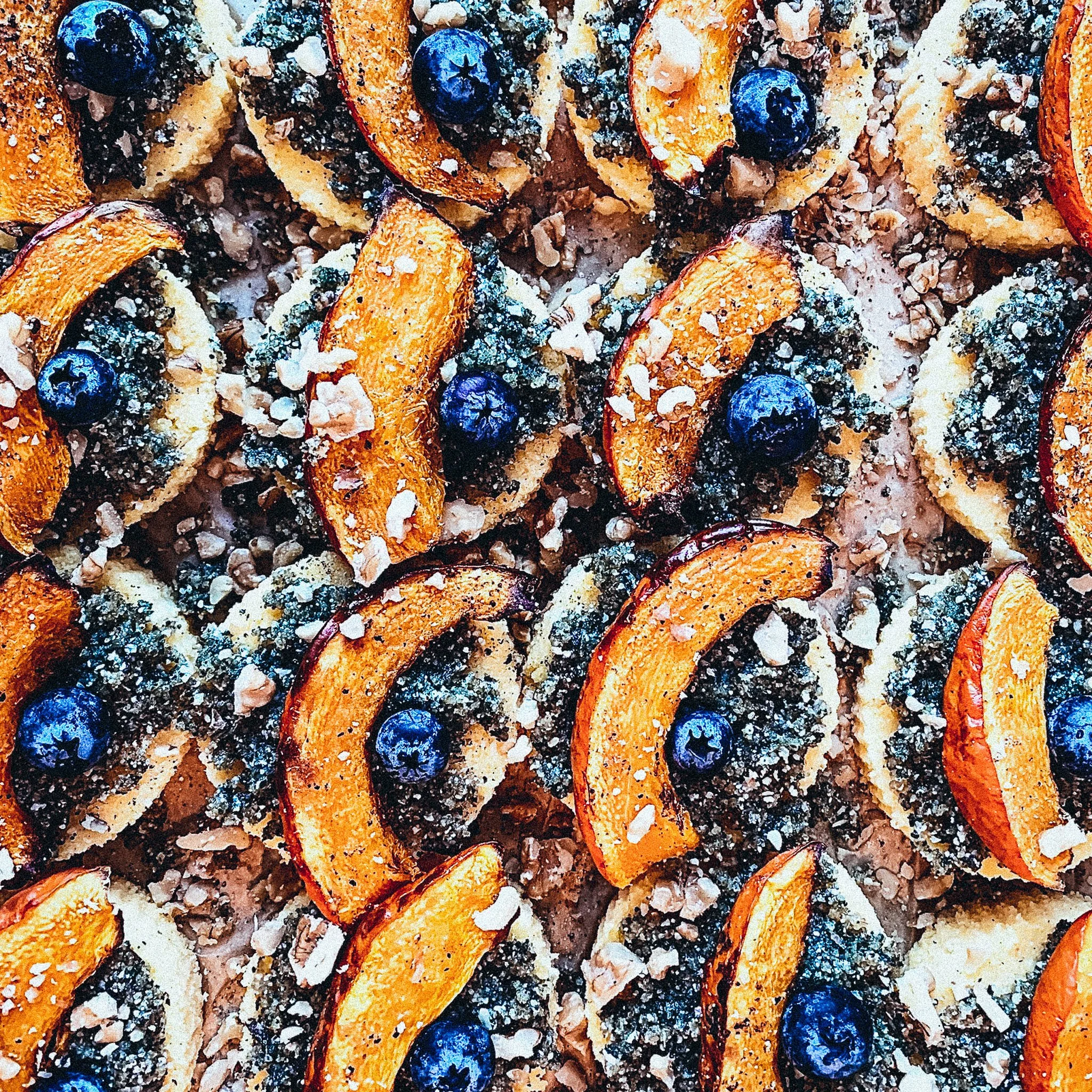 Nahaufnahme von Gebäck oder Keksen, die mit dunkelblauer Füllung, Blaubeeren und orangefarbenen Fruchtscheiben dekoriert sind, möglicherweise Pfirsiche. Es gibt auch Nuss- oder Streuseldekoration auf den Keksen. Die Anordnung ist gleichmäßig und ansprechend gestaltet.