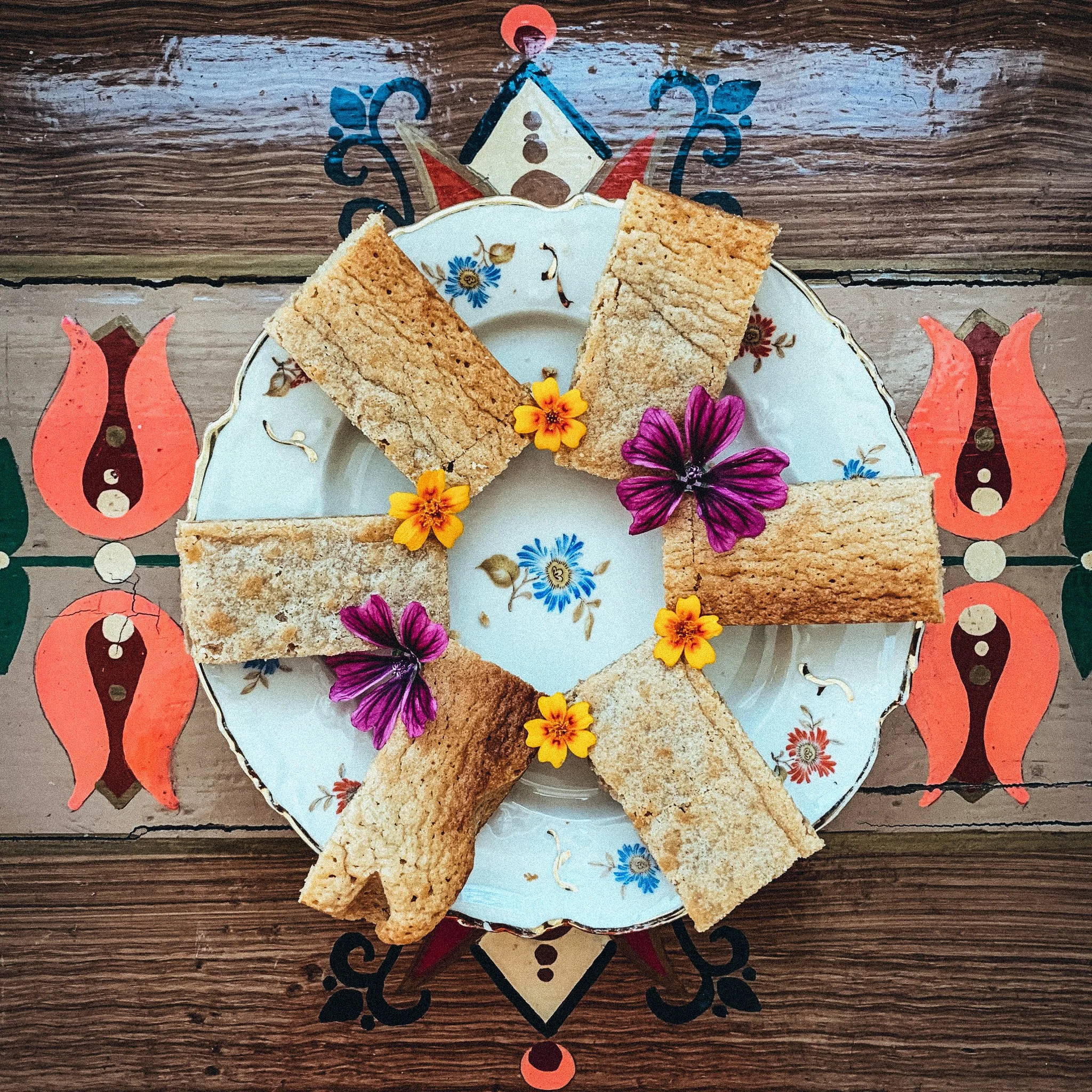 Ein Teller mit rechteckigen Keksen oder Gebäckstücken, dekoriert mit lila und gelben Blumen, auf einem gemusterten Tisch.