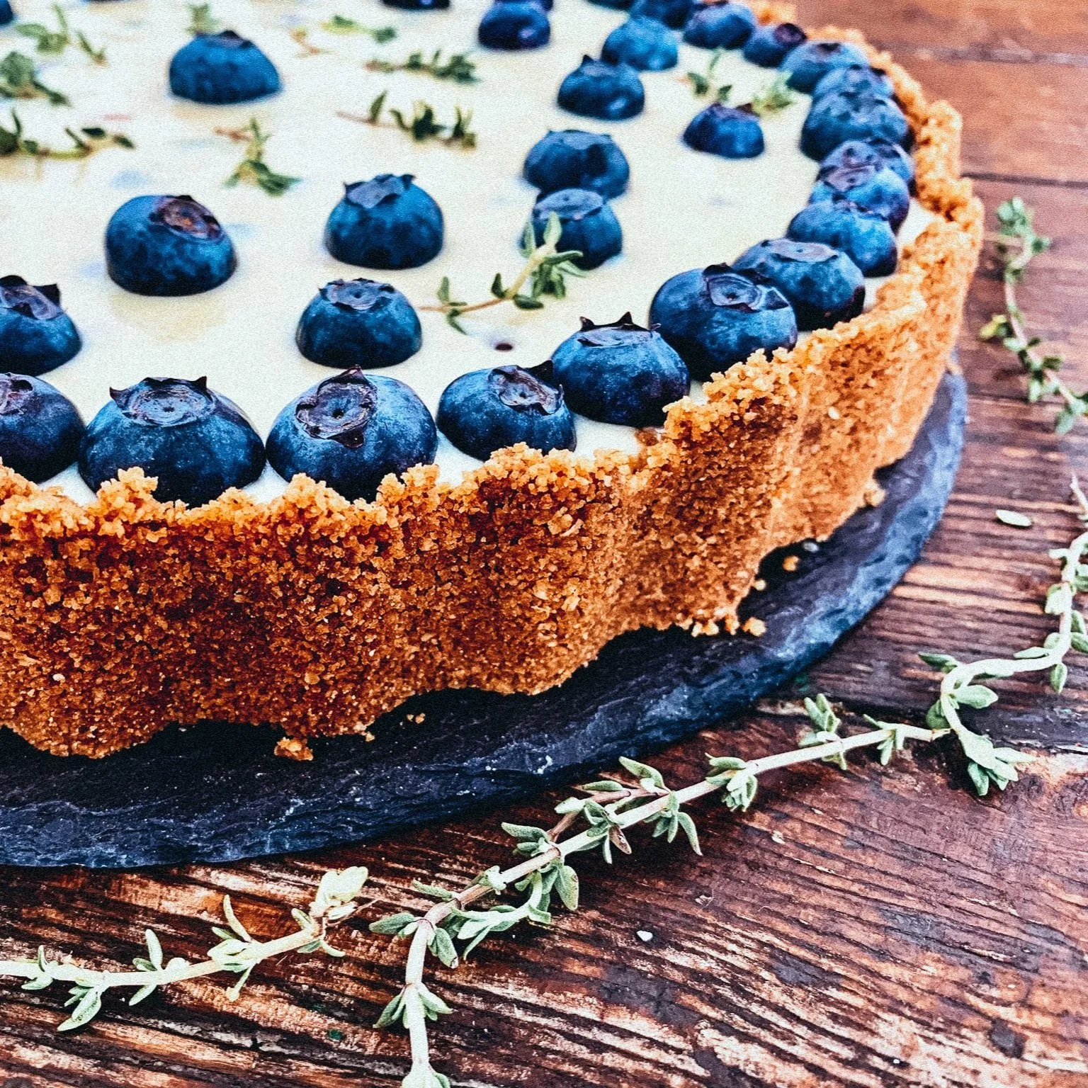 Nahaufnahme eines Heidelbeerkuchens mit Krümelboden, auf einem Holzbrett serviert. Der Kuchen ist mit frischen Heidelbeeren und Kräutern garniert.