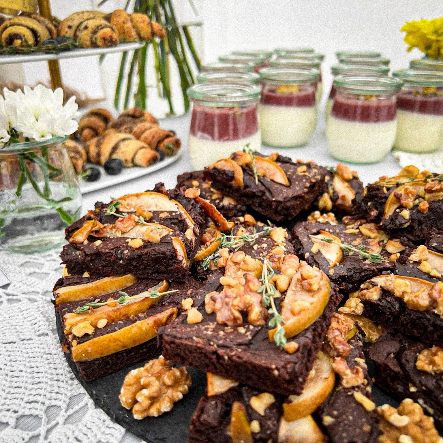 Desserttisch mit Brownies, Nussscheiben, kleinen Gebäckrollen und Gläsern mit Schichtdesserts. Im Hintergrund Blumendekoration auf einer Spitzentischdecke.
