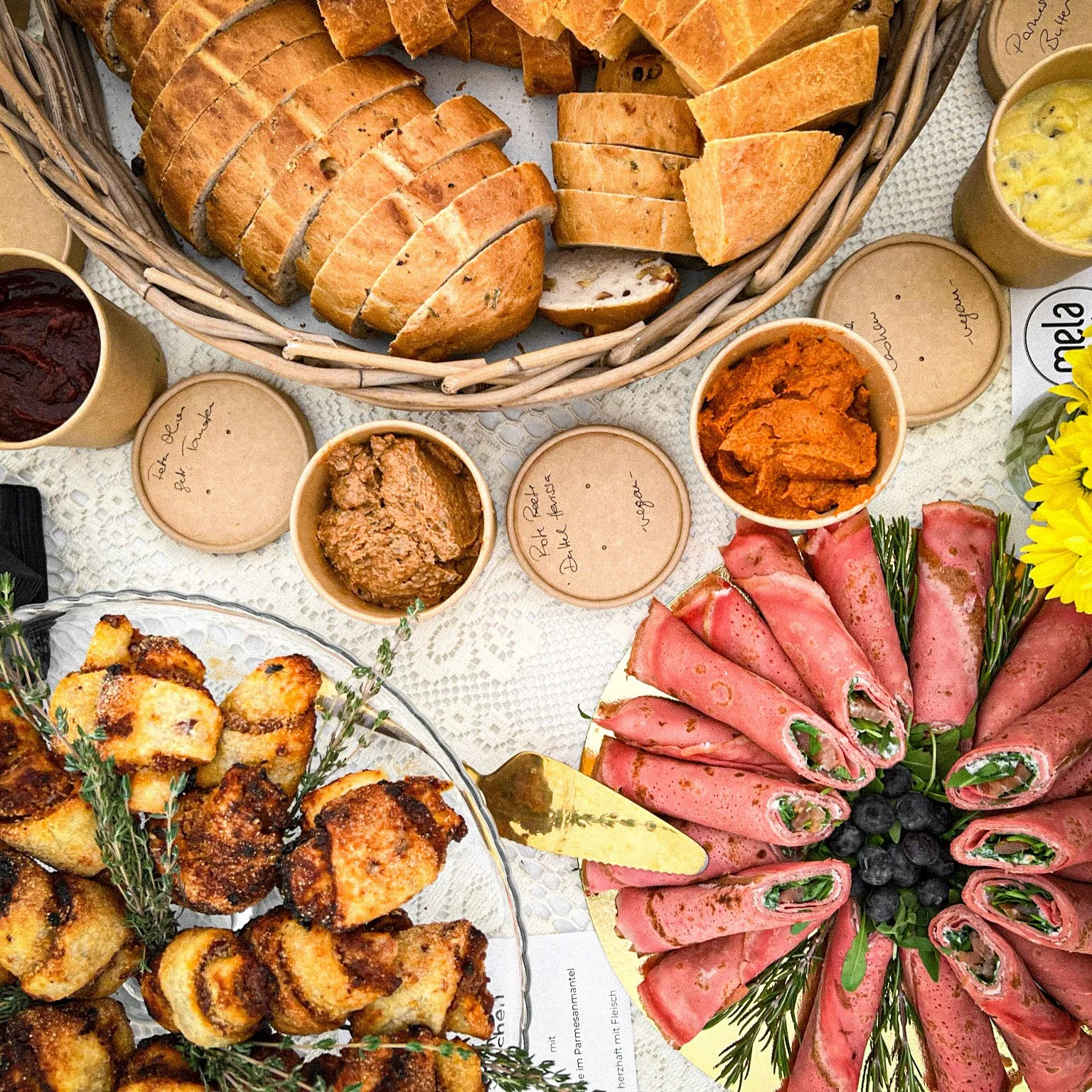Ein Buffet mit verschiedenen Speisen, darunter aufgeschnittenes Brot in einem Korb, mehrere Dips in Schüsseln, gerollte Fleischscheiben mit Füllung, gebackene Teigteilchen auf einem Teller, und Dekorationen mit frischen Kräutern. Alles auf einem hellen Tischdecke präsentiert.