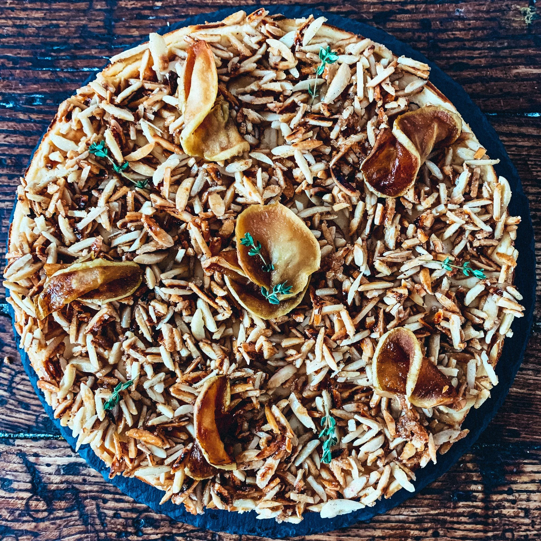 Ein runder Kuchen mit einer knusprigen, gebräunten Haube aus gerösteten Mandelstiften, dekoriert mit getrockneten Apfelscheiben und Kräutern auf einem dunklen Teller auf Holztisch.