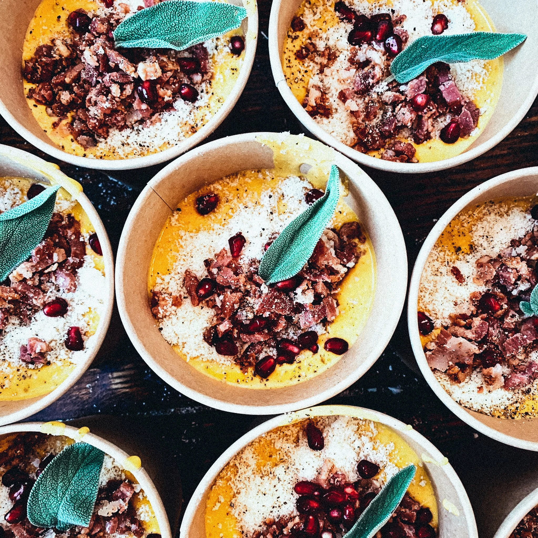 Mehrere Schüsseln mit Polenta, Granatapfelkernen, Speckstücken und einem grünen Blatt als Garnierung, von oben fotografiert.