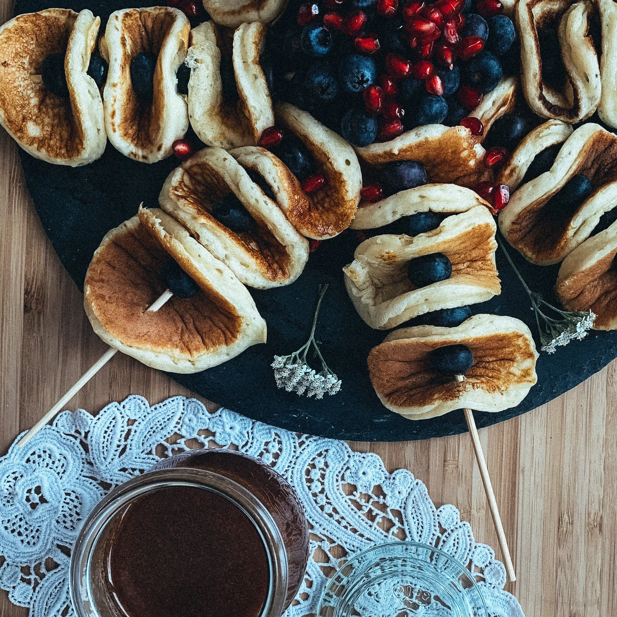 Mini-Pfannkuchen auf Holzspießen, garniert mit Heidelbeeren und Granatapfelkernen, auf einem dunklen Servierbrett auf Spitze, mit einem Glas Schokoladensauce daneben.