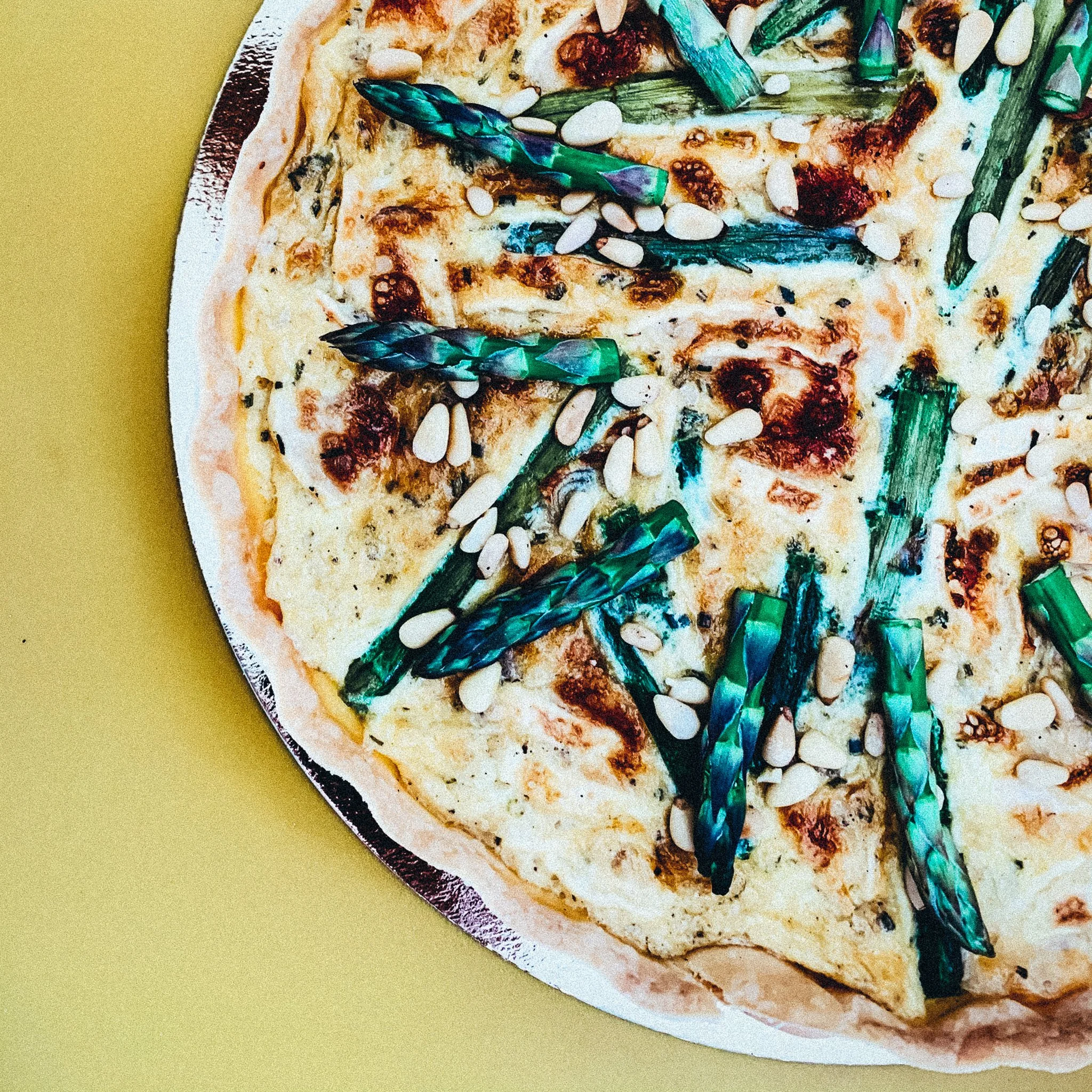 Pizza mit grünem Spargel und Pinienkernen auf gelbem Hintergrund.