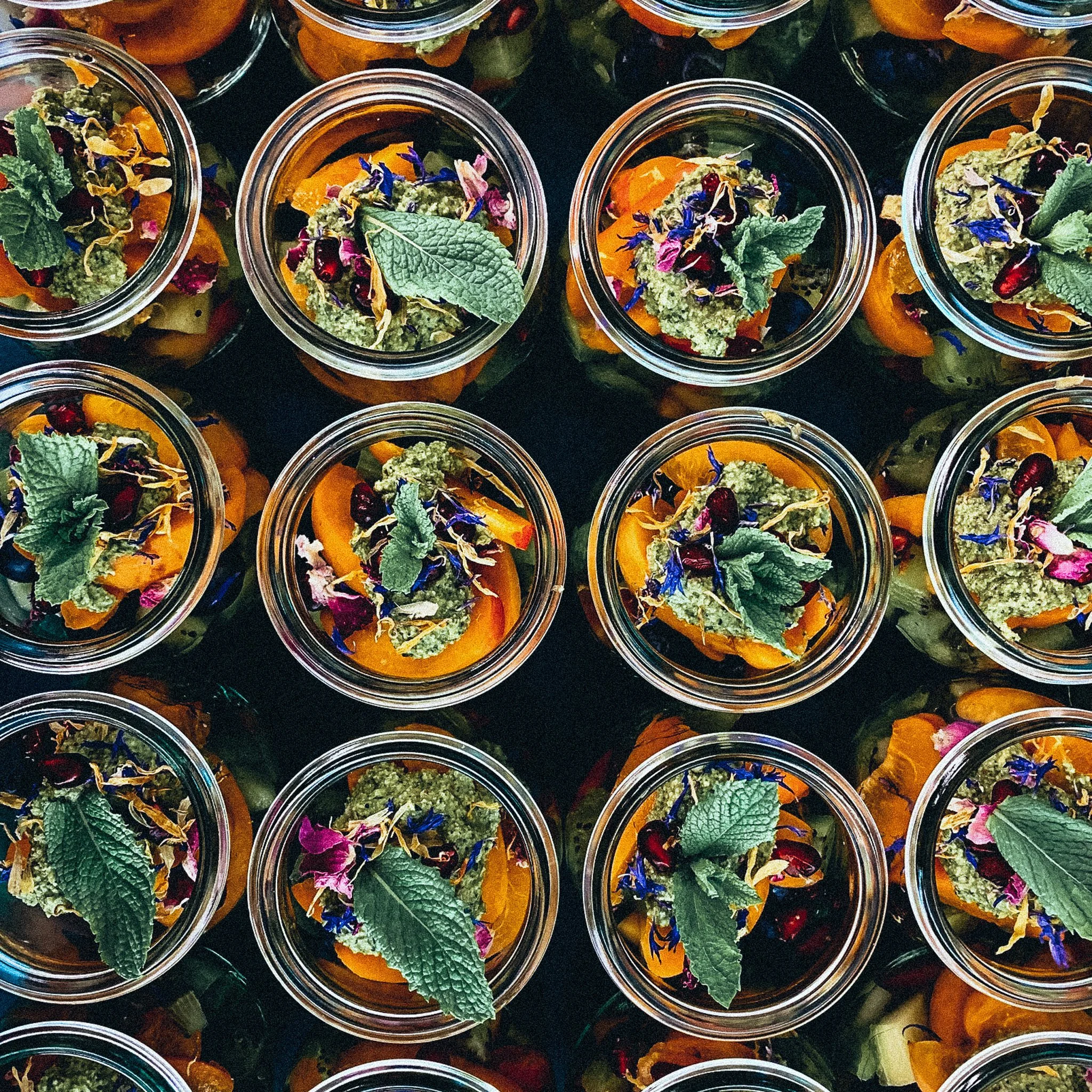 Mehrere Gläser mit buntem, geschichtetem Essen, garniert mit frischen Kräutern und essbaren Blüten.