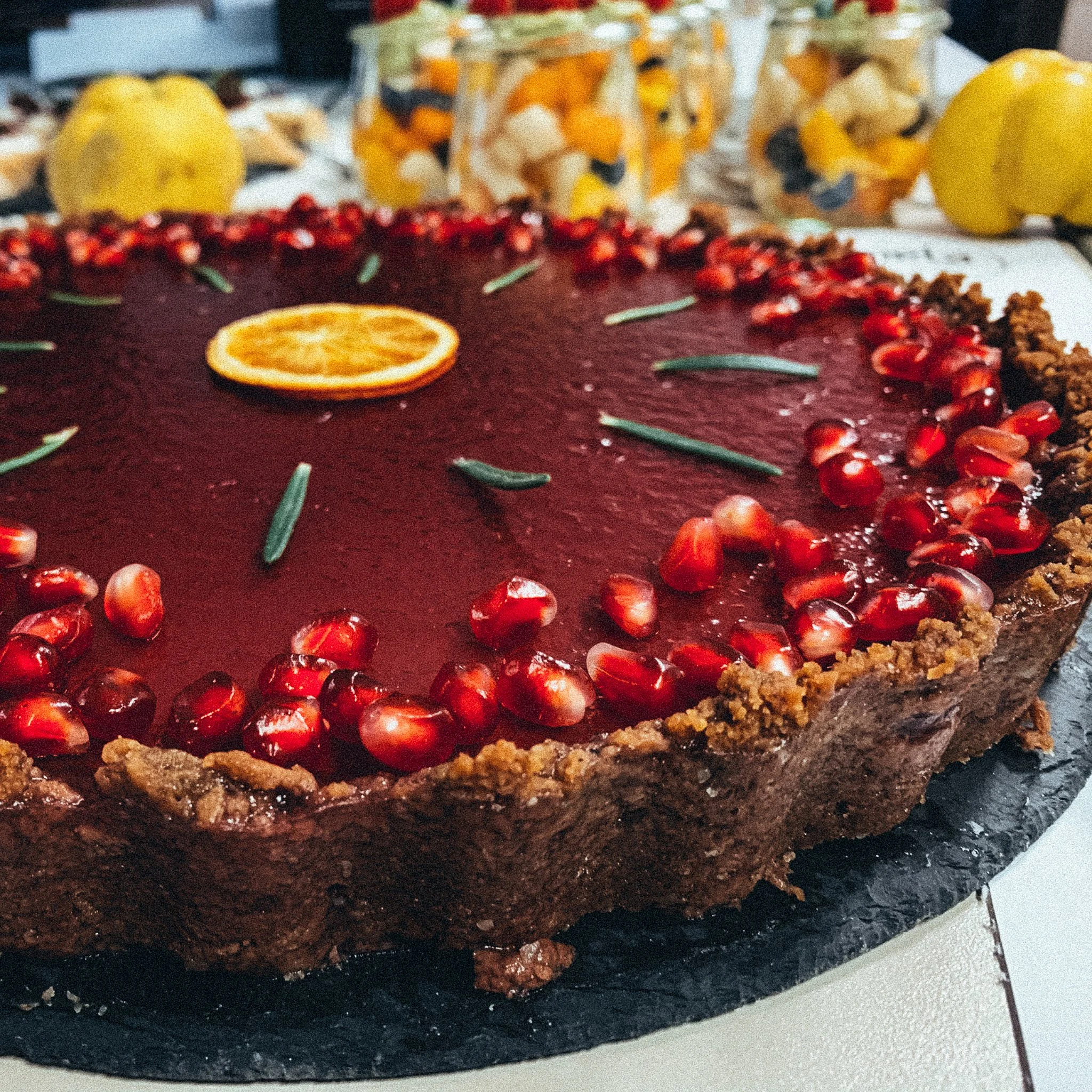 Nahaufnahme einer Torte mit roter Glasur, dekoriert mit Granatapfelkernen, Rosmarin und einer Orangen-Scheibe. Im Hintergrund sind gelbe Früchte und Glasbehälter mit Obst zu sehen.